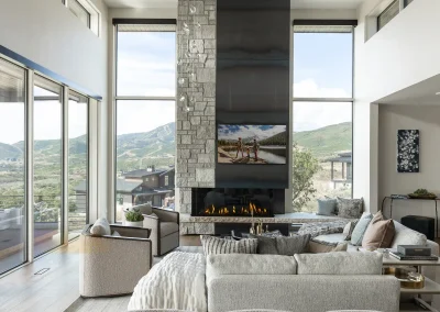 Modern living room with floor-to-ceiling windows overlooking mountains, featuring a stone fireplace with a mounted TV, neutral-toned seating, and abundant natural light.