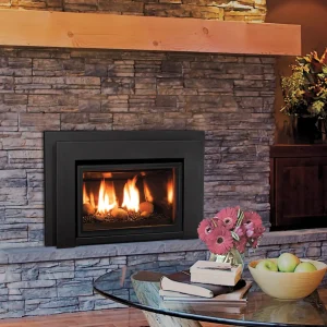 Enviro E20 gas fireplace insert with black frame set in a stacked stone wall, featuring warm flames, a wooden mantel, and a cozy living room with flowers and a bowl of green apples on a glass coffee table.