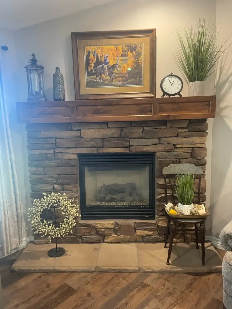 Rustic stone fireplace with a thick wooden mantel, decorated with framed artwork, lanterns, a clock, and potted greenery in a cozy living space.