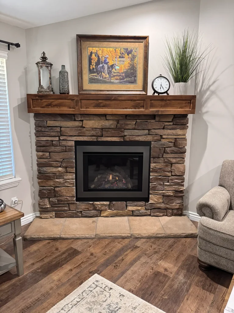 Rustic stone fireplace with a thick wooden mantel, decorated with framed artwork, lanterns, a clock, and potted greenery in a cozy living space.